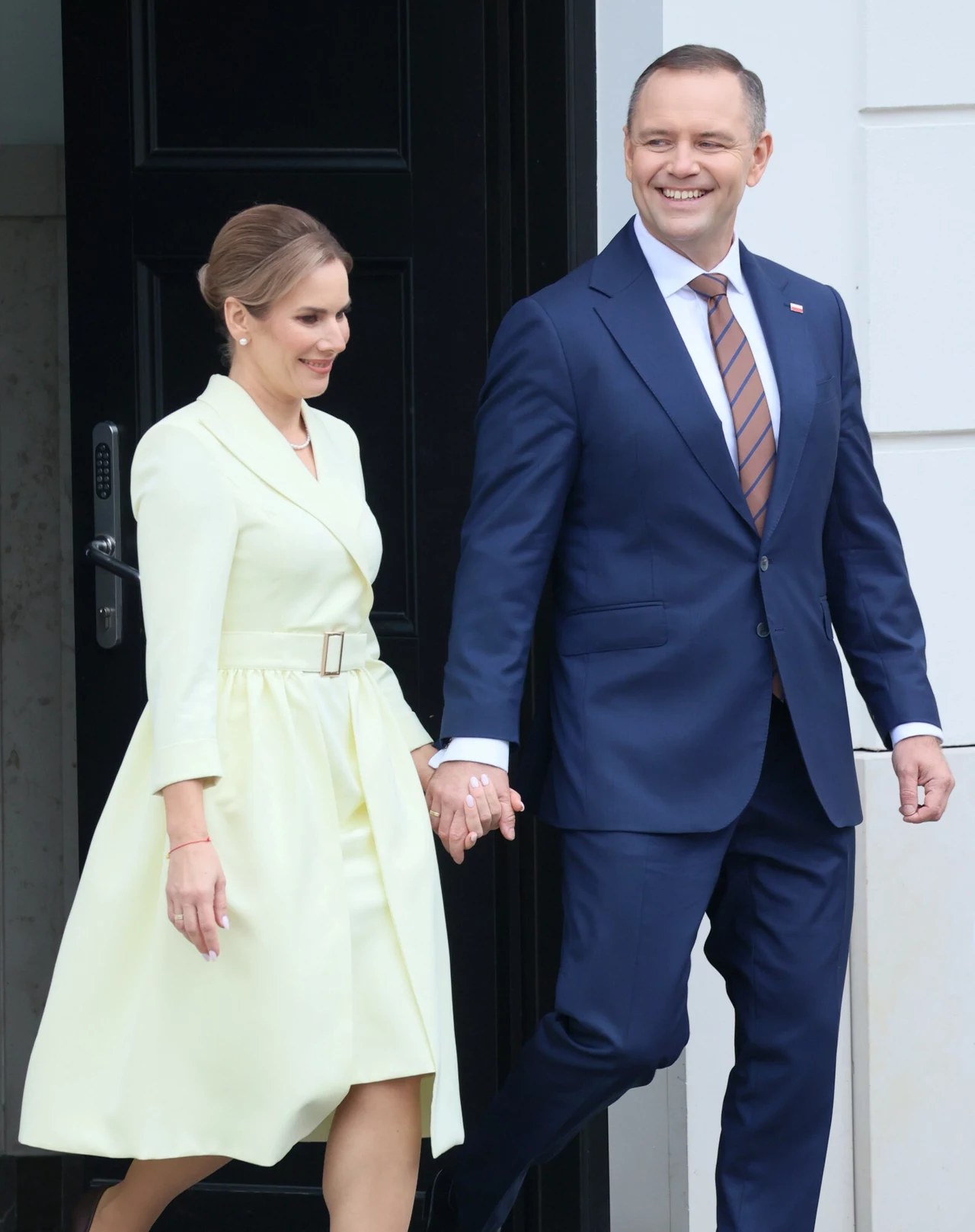 Elegancko ubrana para trzymająca się za ręce, opuszczająca budynek. Kobieta w jasnożółtej sukience, mężczyzna w granatowym garniturze z krawatem w paski, oboje uśmiechnięci i wyglądający na zadowolonych.