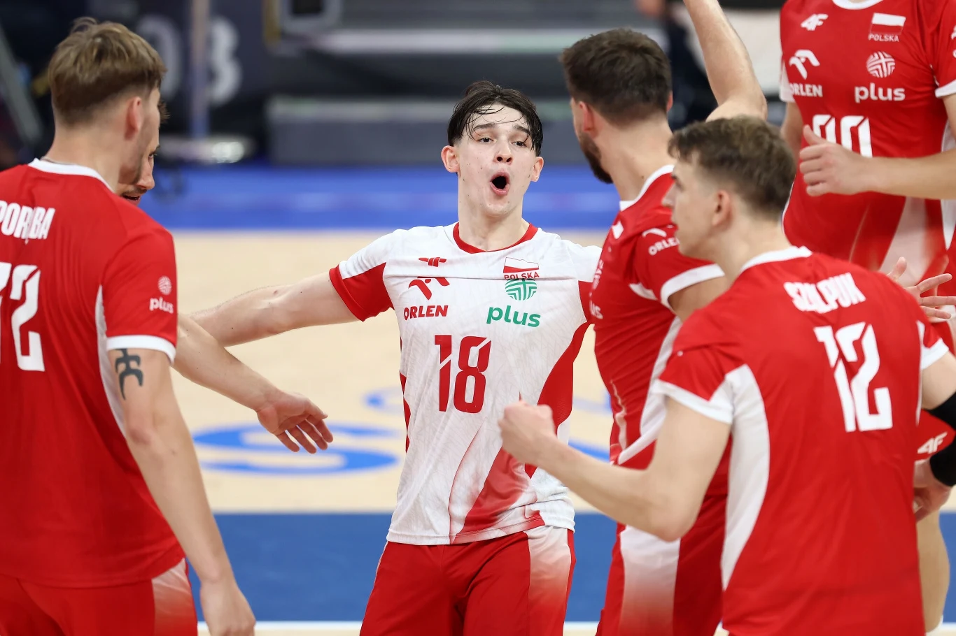 Grupa siatkarzy w czerwono-białych strojach podczas dynamicznej celebracji punktu na boisku, zawodnik ze środkowym numerem 18 wyraża silne emocje, gestykulując w kierunku kolegów z drużyny.