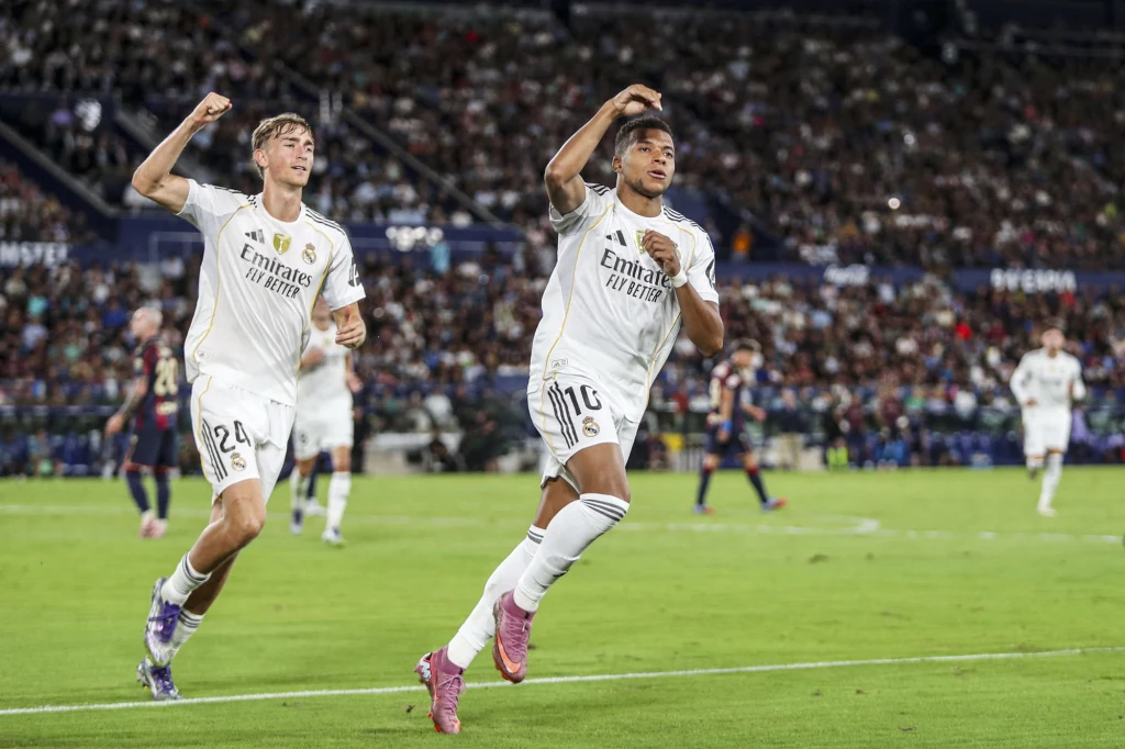 Dejan Huijsen i Kylian Mbappe w barwach Realu Madryt Dwoje piłkarzy w białych strojach świętuje zdobycie bramki na murawie stadionu, w tle widać kibiców na trybunach i kilku zawodników przeciwnika.