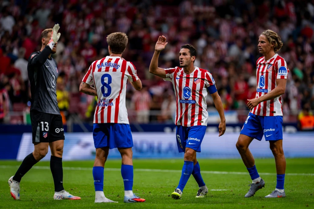 Piłkarze Atletico Madryt w meczu LA Liga z Villarrealem Piłkarze drużyny w czerwono-białych pasiastych koszulkach oraz niebieskich spodenkach znajdują się na murawie stadionu, wzywając się nawzajem lub świętując, w tle rozmyty tłum kibiców.