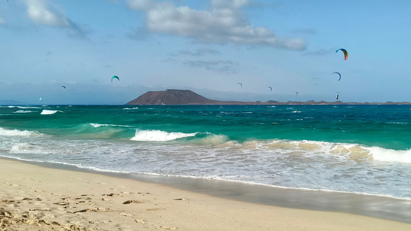 Wyspa Lobos widziana z Fuerteventury Piaszczysta plaża z turkusowymi falami oceanu oraz kilkoma kolorowymi latawcami kite surferów unoszącymi się nad wodą, w tle wyłania się wyspa o płaskim szczycie pod rozproszonymi chmurami.