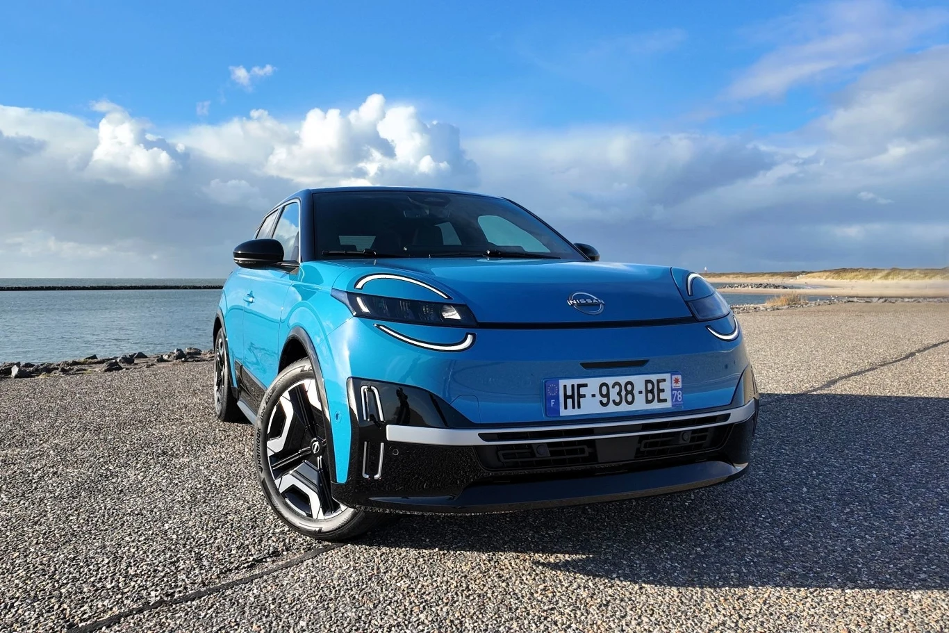 Nissan Micra powraca po kilku latach nieobecności. Teraz jest elektrykiem. Niebieski samochód terenowy stoi zaparkowany na asfaltowej nawierzchni przy wybrzeżu pod lekko zachmurzonym niebem, w tle widoczna woda oraz fragment plaży.