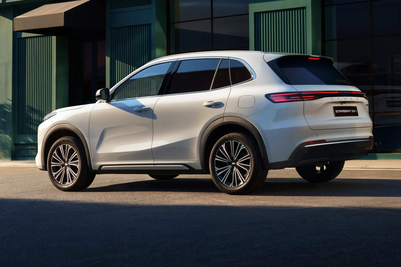 Biały nowoczesny SUV zaparkowany na ulicy, w tle zielone fasady budynków i przeszklenia, samochód w pełnym słońcu widoczny z tyłu oraz z boku.