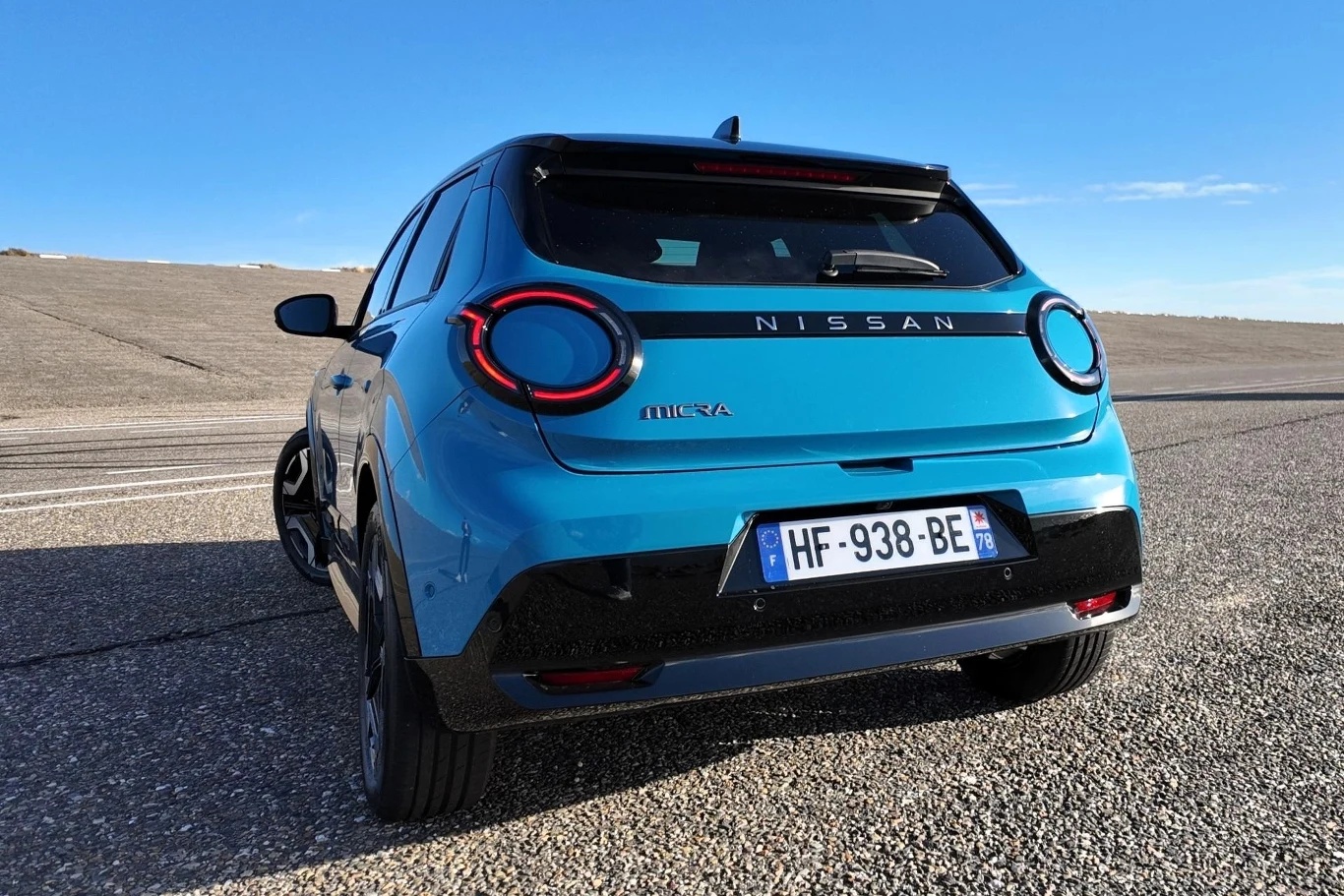 Okrągłe światła z przodu i z tyłu optycznie łączą się z wyżłobieniem biegnącym po bokach samochodu. Nowoczesny niebieski samochód Nissan Micra na opustoszałej nawierzchni drogowej, tył pojazdu z wyraźnie widocznymi światłami i tablicą rejestracyjną na tle nieba.