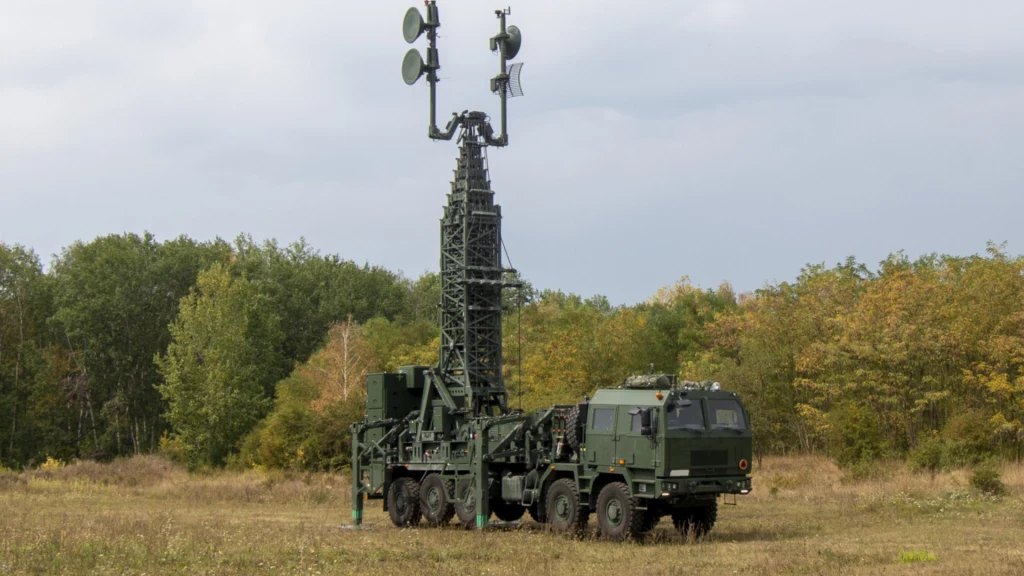 Wojskowy pojazd specjalistyczny z zainstalowaną wysoką wieżą antenową na otwartym terenie, w tle drzewa i niebo.