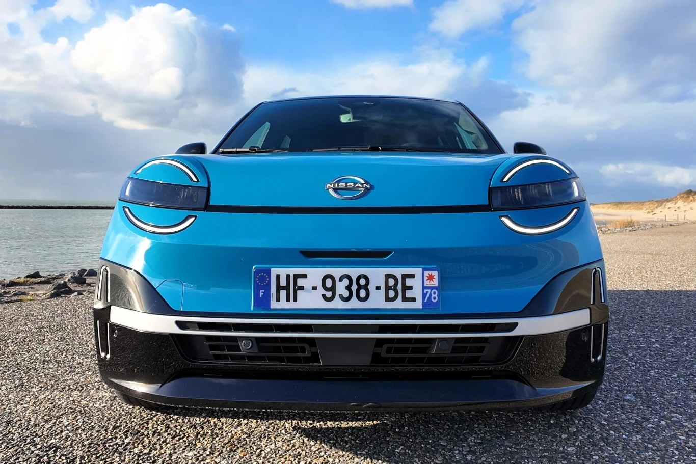 Na jednym ładowaniu Nissan Micra ma przejechać do 416 km. Nowoczesny, niebieski samochód elektryczny marki Nissan zaparkowany na otwartym terenie przy wodzie, widok od frontu z wyraźnie widoczną tablicą rejestracyjną i charakterystycznym wzornictwem świateł.