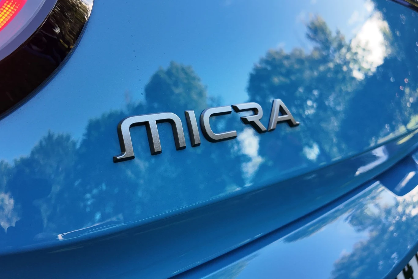 Ceny nowej Micry zaczynają się od 124 900 zł. Niebieski lakier samochodu z wyeksponowanym napisem 'Micra'; na karoserii odbijają się drzewa i niebo