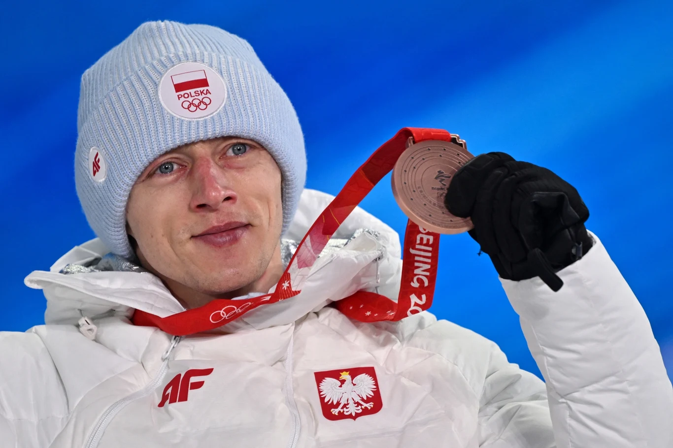 Dawid Kubacki z brązowym medalem igrzysk olimpijskich Sportowiec ubrany w białą kurtkę i błękitną czapkę z logo Polskiego Komitetu Olimpijskiego trzyma brązowy medal olimpijski, w tle niebieskie rozmycie.