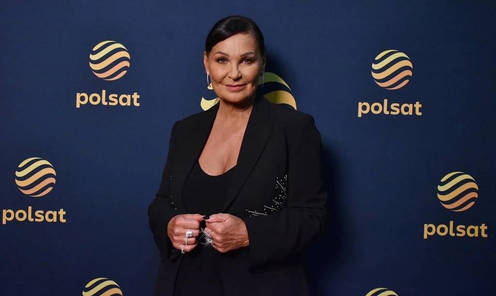 Kobieta w eleganckim czarnym stroju na tle ścianki z logotypami Polsat, uśmiechnięta, z ciemnymi włosami upiętymi do tyłu, ubrana w czarną marynarkę, stoi z dłońmi splecionymi na wysokości talii