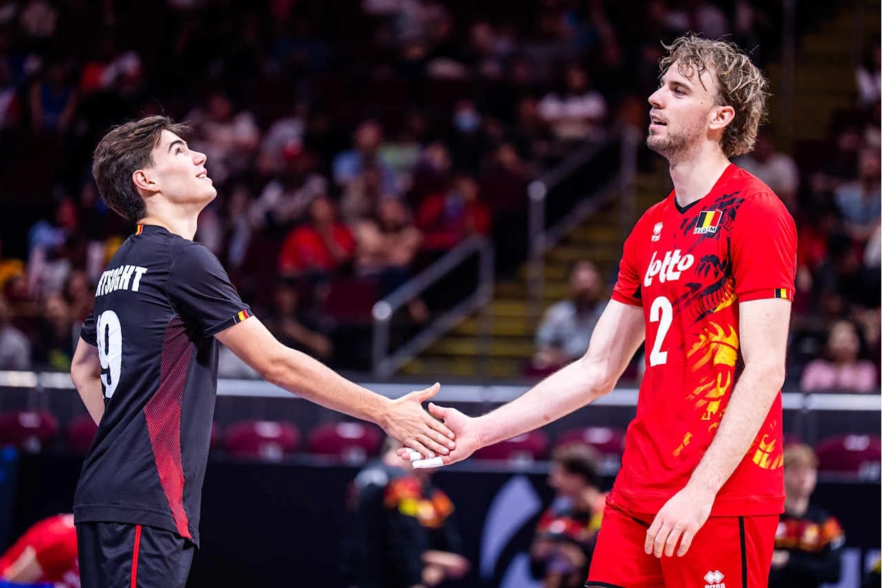 Siatkarze reprezentacji Belgii: Gorik Lantsoght i Ferre Reggers Dwóch siatkarzy w sportowych strojach ściska sobie dłonie na tle trybun, jeden z nich ubrany w czerwony strój z numerem 2, drugi w ciemniejszym stroju z numerem 9, wokół nich widoczni kibice na widowni.