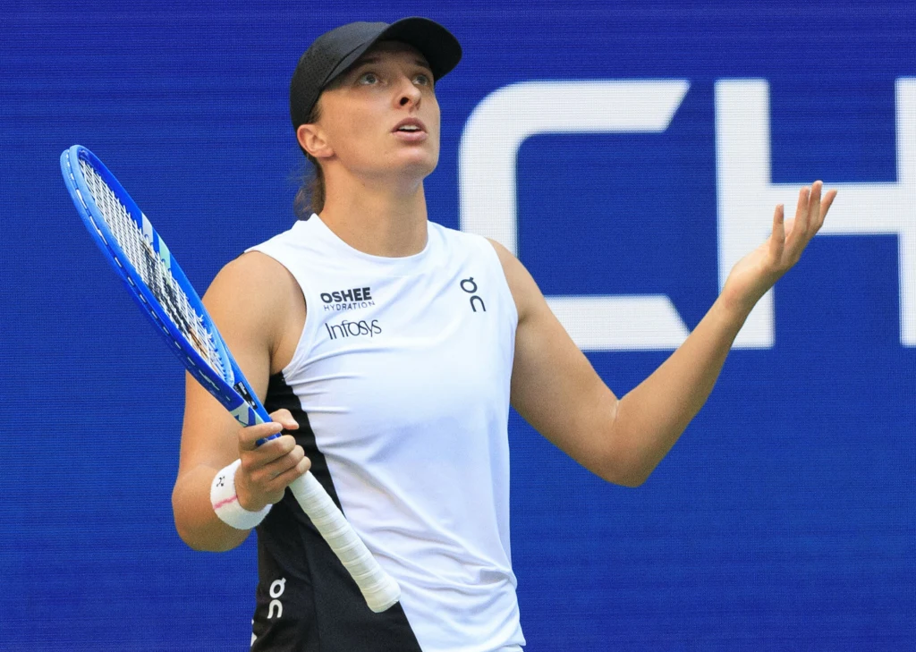 Tenisistka ubrana w biały strój sportowy i czarną czapkę trzyma w lewej ręce rakietę, stojąc na tle niebieskiej kortowej ściany z fragmentem napisu. Jej mina i rozłożone ręce wyrażają zdziwienie lub zawód.