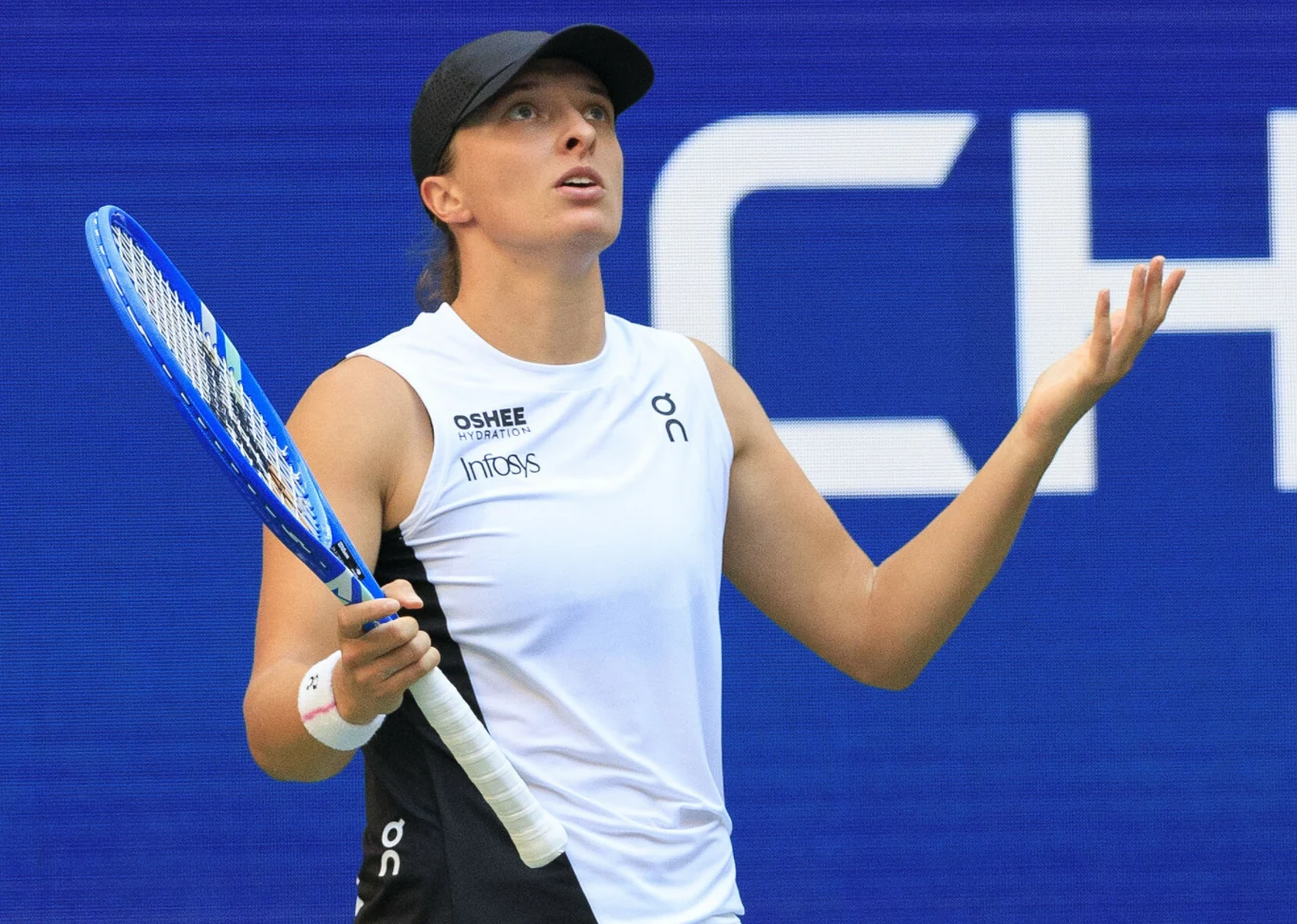 Tenisistka ubrana w biały strój sportowy i czarną czapkę trzyma w lewej ręce rakietę, stojąc na tle niebieskiej kortowej ściany z fragmentem napisu. Jej mina i rozłożone ręce wyrażają zdziwienie lub zawód.