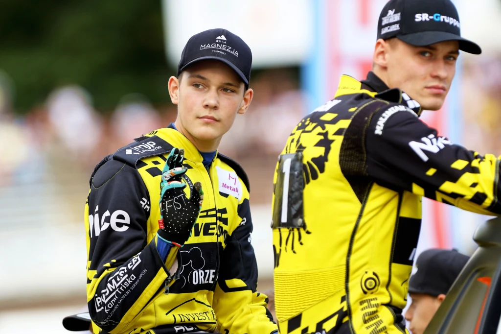 Dwóch młodych żużlowców w jaskrawo żółto-czarnych kombinezonach, jeden z nich spogląda w stronę obiektywu i unosi rękę w geście pozdrowienia, tło nieostre, publiczność zamazana.