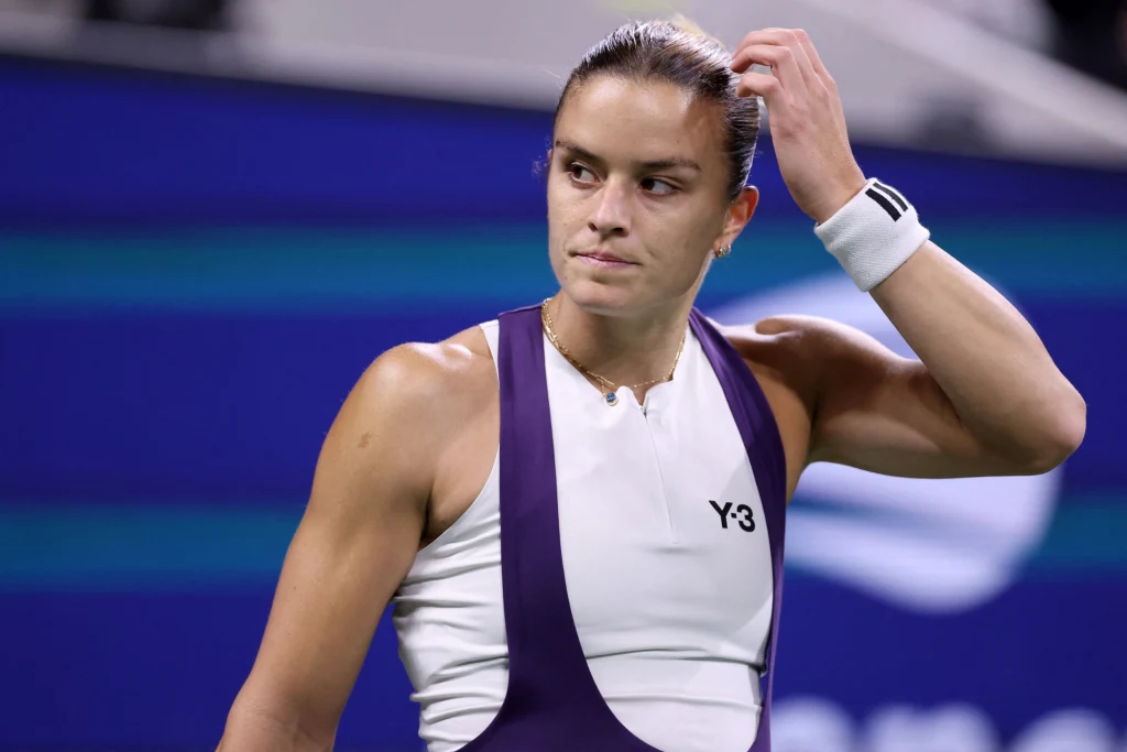 Maria Sakkari Tenisistka w sportowej stylizacji w trakcie meczu na profesjonalnym korcie, poprawia włosy, sylwetka wyraźnie zarysowana na tle niebieskich trybun.