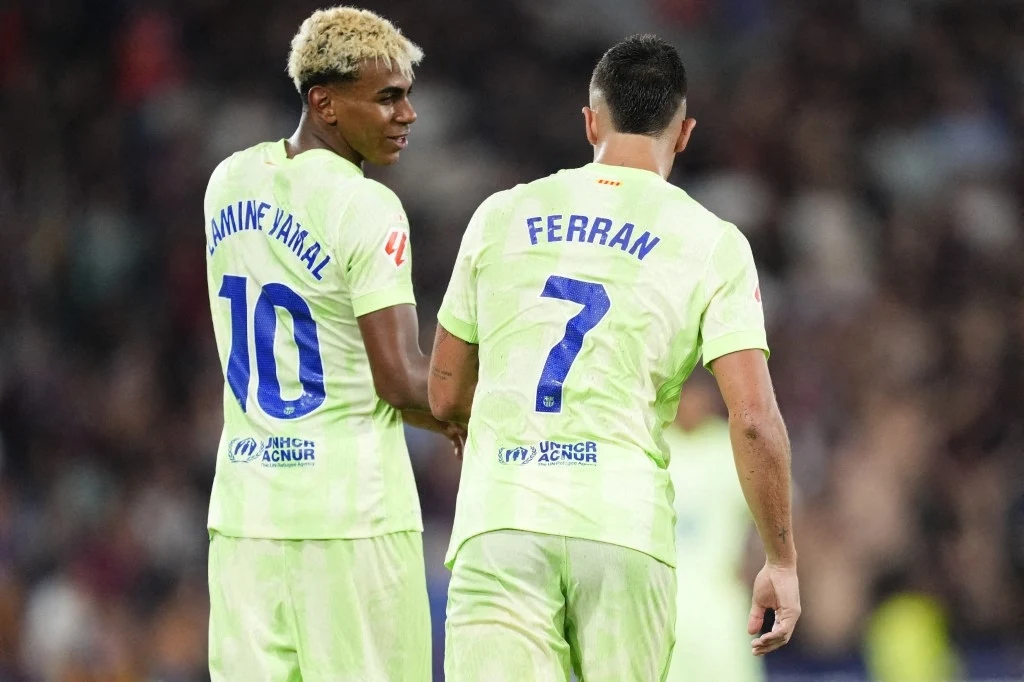 Lamine Yamal wraca do zdrowia Dwóch piłkarzy drużyny w jasnozielonych strojach z widocznymi numerami 10 i 7 idzie razem po murawie, prowadząc rozmowę. Stadion w tle jest rozmyty, podkreślając sportową atmosferę.