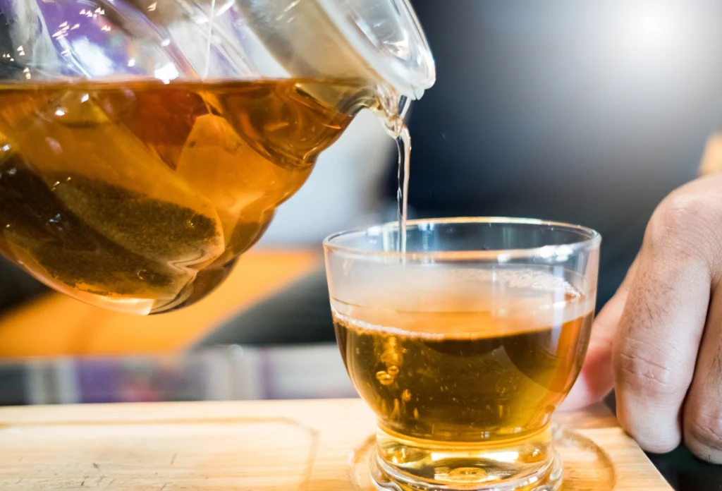 Napary ziołowe działają dobroczynnie na cały organizm. Wspierają również odporność Szklanka napełniana gorącą herbatą z przezroczystego dzbanka trzymanego nad filiżanką, w tle widoczna ręka chwytająca kubek, całość ustawiona na drewnianej powierzchni, delikatne rozmycie tła.