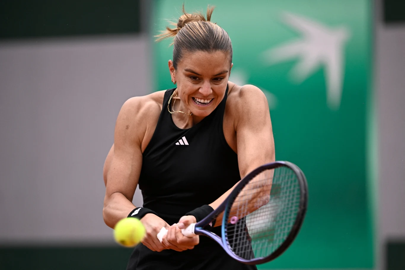 Zawodniczka podczas intensywnego uderzenia dwuręcznym backhandem tenisowym, skupiona twarz, sportowy strój, lotka piłki nad rakietą, dynamiczny ruch i tło z zielonym elementem graficznym.