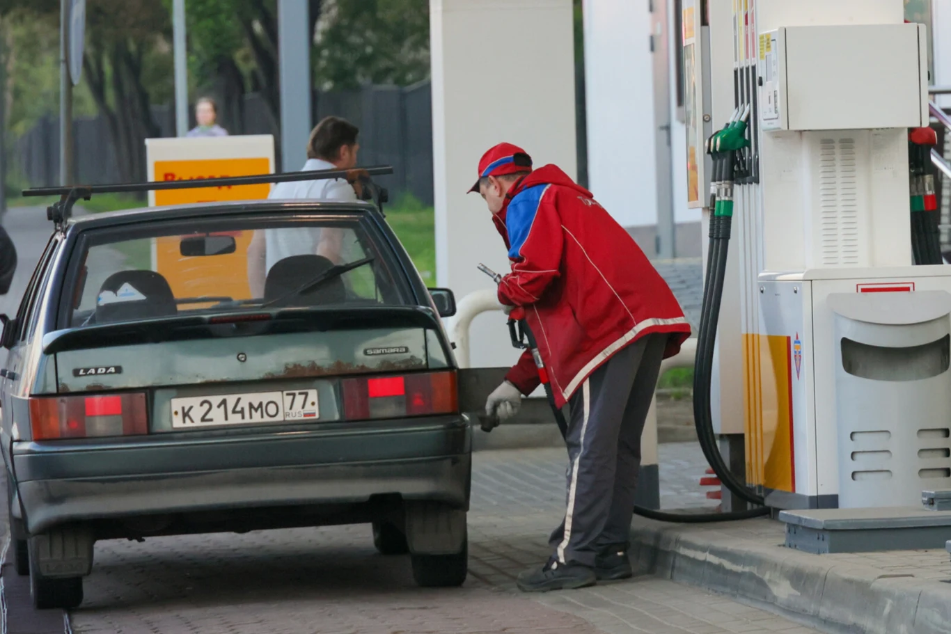 Ataki dronów na rosyjską infrastrukturę. Zaczyna brakować paliwa Pracownik stacji benzynowej w czerwonej kurtce tankuje samochód osobowy marki Lada przy dystrybutorze paliwa, w tle widoczna kobieta oraz elementy stacji.