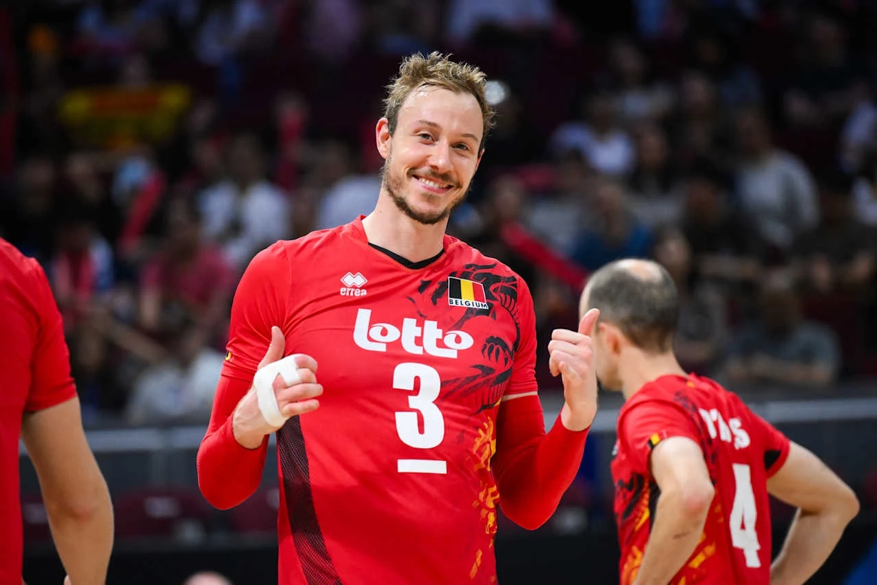 Siatkarski zawodnik w czerwonym stroju z numerem 3 i belgijską flagą uśmiecha się i pokazuje gest kciuków do góry, obok niego stoi inny siatkarz z numerem 4 odwrócony tyłem, w tle widownia sportowa.