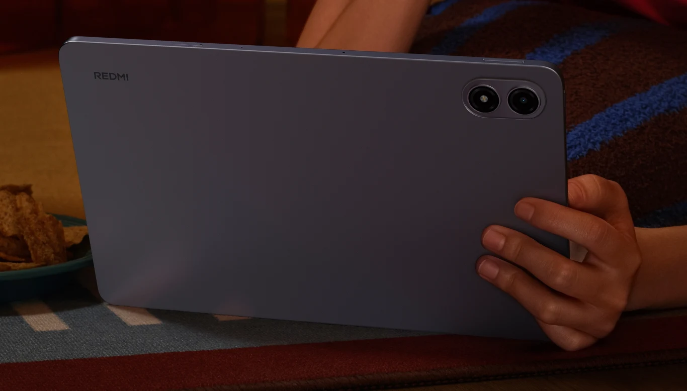Osoba trzyma wysokiej klasy tablet marki Redmi w poziomej pozycji, w tle koc z kolorowymi paskami oraz miseczka z przekąskami po lewej stronie kadru.