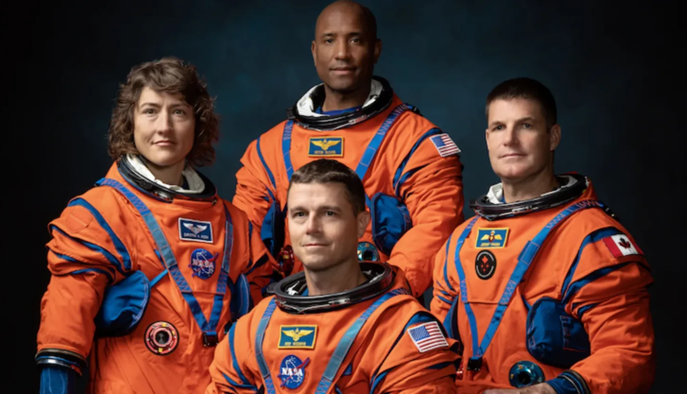 Załoga misji Artemis II (od lewej): Christina Koch, Victor Glover (pilot), Reid Wiseman (dowódca) i Jeremy Hansen Cztery osoby w pomarańczowych skafandrach kosmicznych z widocznymi naszywkami NASA oraz flagami państw, ustawione w szeregu na neutralnym, ciemnym tle, tworzą oficjalny portret załogi.