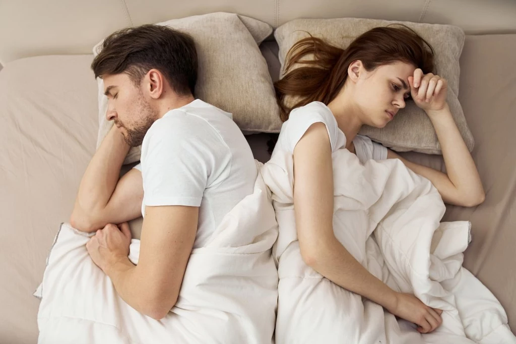 Para leży w łóżku tyłem do siebie, oboje mają niezadowolone miny, każde spogląda w inną stronę, co sugeruje napiętą atmosferę lub konflikt emocjonalny.