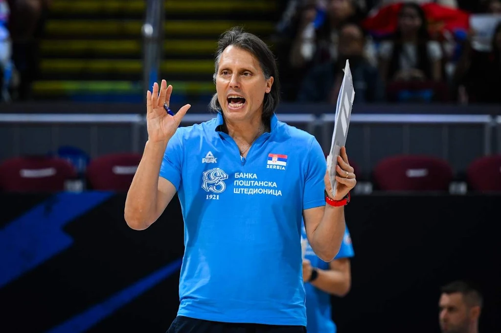 MŚ siatkarzy. Gheorghe Cretu, trener reprezentacji Serbii Trener prowadzi drużynę sportową podczas meczu, ubrany w niebieską koszulkę z logotypem klubu, trzyma w lewej dłoni tabliczkę i energicznie gestykuluje, w tle widoczna widownia.