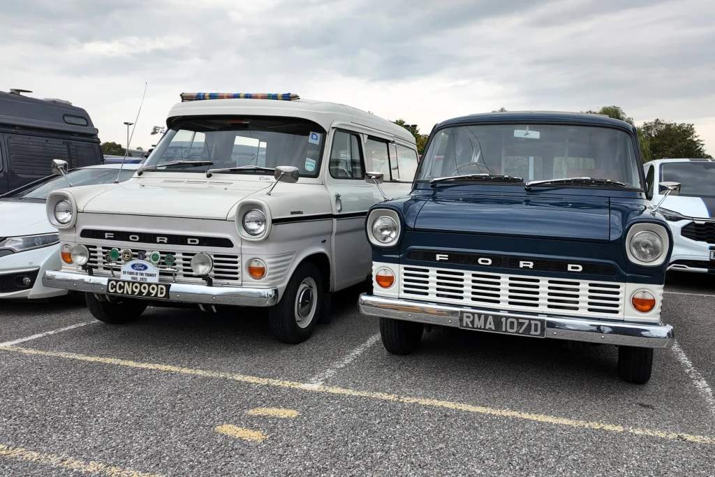 Dwa zabytkowe samochody marki Ford zaparkowane obok siebie na asfaltowym parkingu, po lewej biały pojazd z lampami ostrzegawczymi na dachu, po prawej ciemnoniebieski samochód o klasycznym wyglądzie, oba otoczone innymi pojazdami.