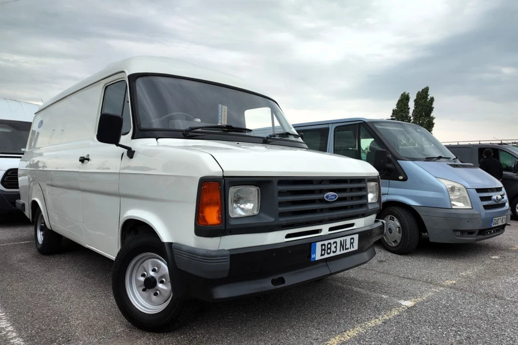 Dwa białe vany marki Ford stojące na parkingu, jeden z nich to starszy model, drugi nowszy, tło stanowią zaparkowane pojazdy oraz pochmurne niebo.
