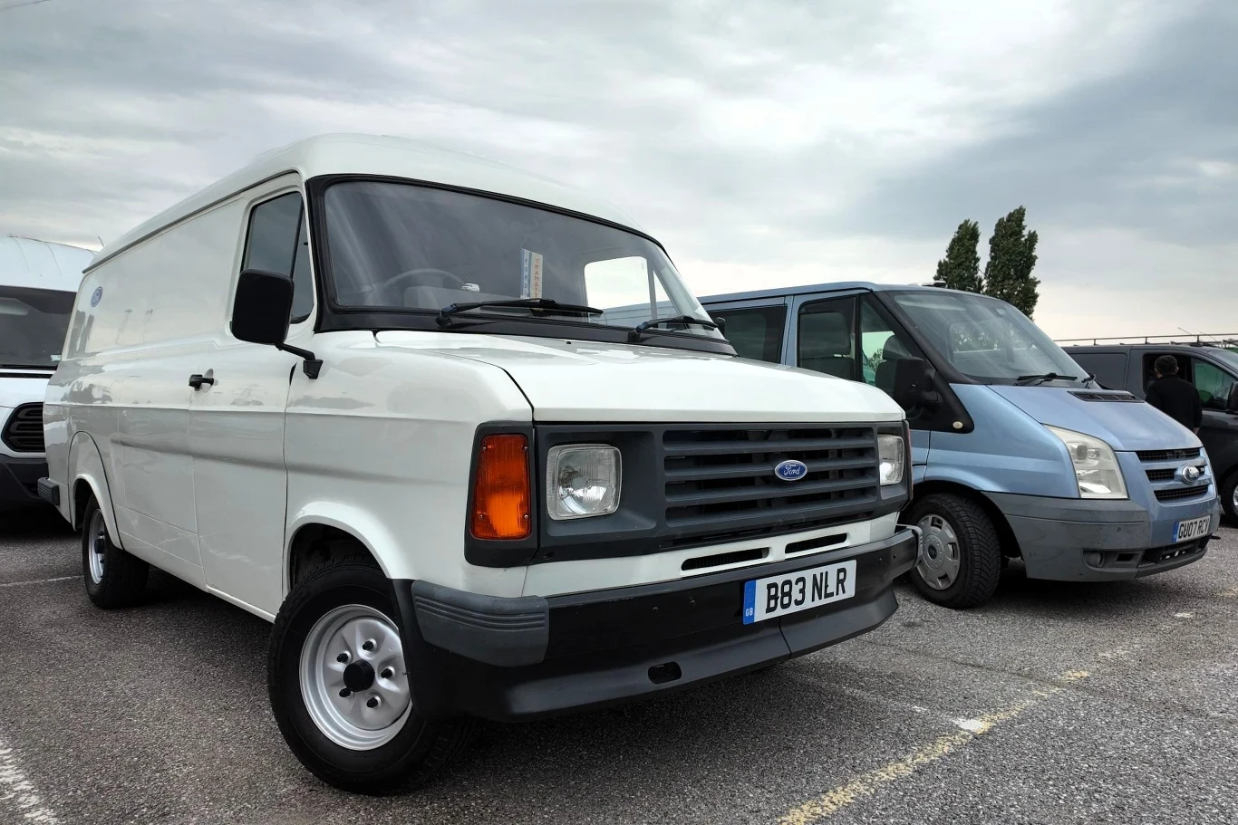 Dwa białe vany marki Ford stojące na parkingu, jeden z nich to starszy model, drugi nowszy, tło stanowią zaparkowane pojazdy oraz pochmurne niebo.