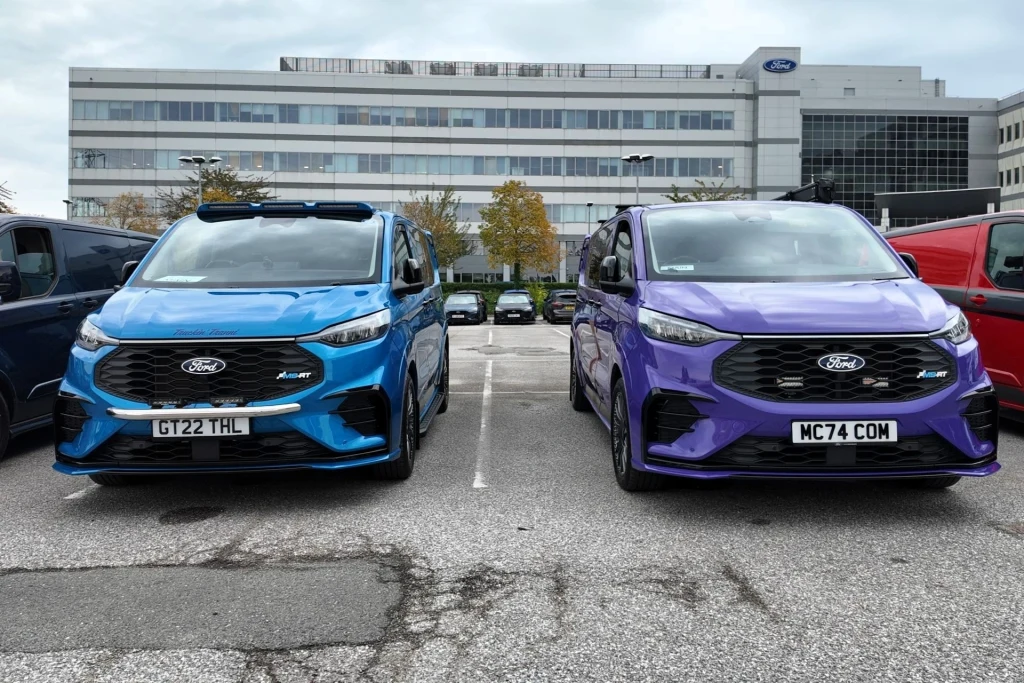 Dwa nowe samochody marki Ford w kolorach niebieskim i fioletowym zaparkowane obok siebie na parkingu przed dużym biurowcem z logo Forda.