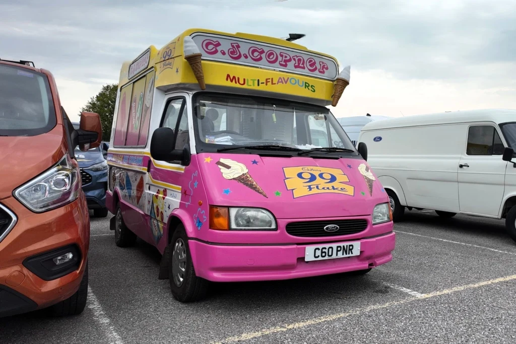 Różowy samochód dostawczy stylizowany na wóz z lodami, ozdobiony kolorową grafiką lodów oraz napisami reklamowymi, zaparkowany na parkingu między innymi samochodami.