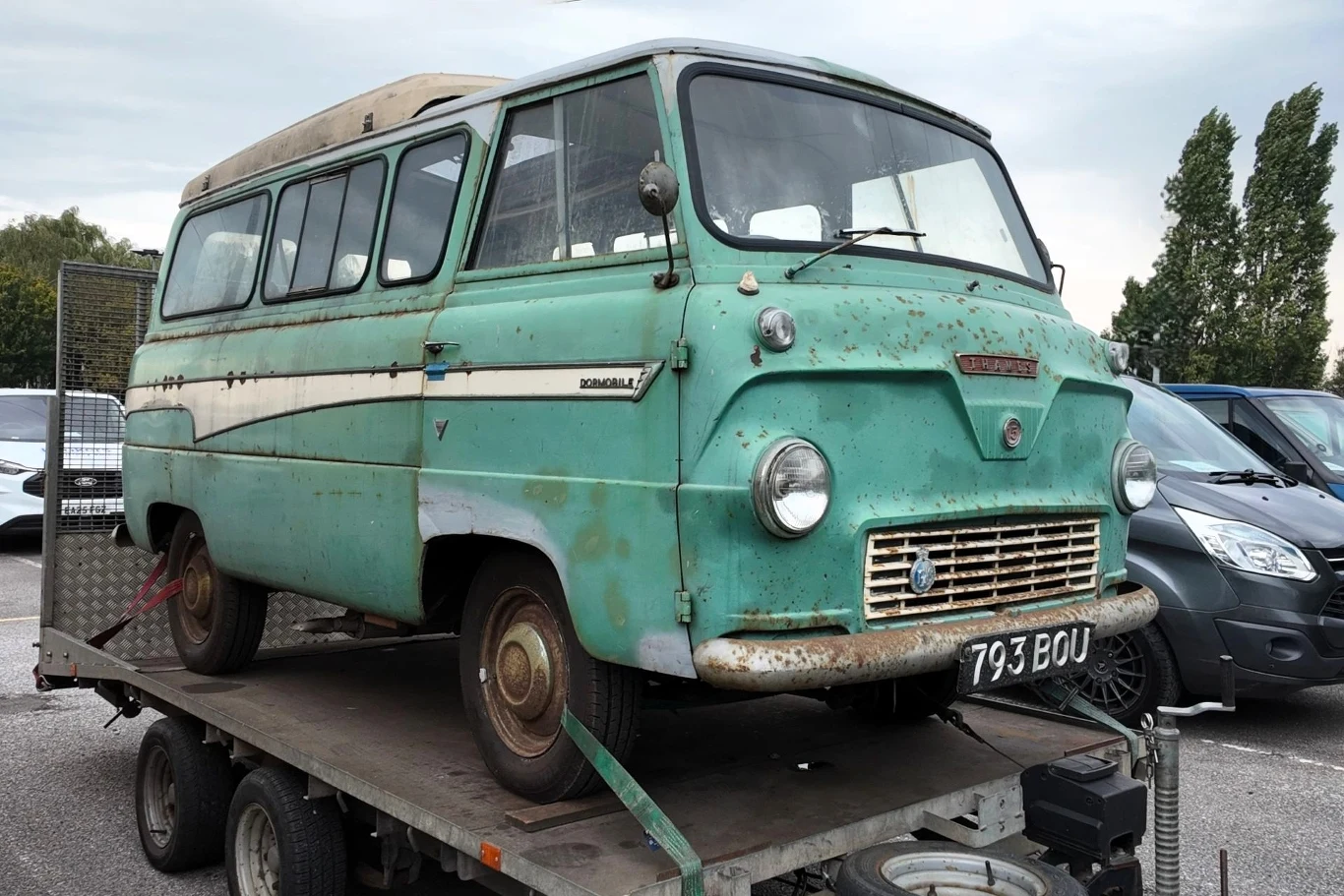 Stary zielony bus marki Bedford z widocznymi śladami rdzy stoi na przyczepie na parkingu, obok innych samochodów.