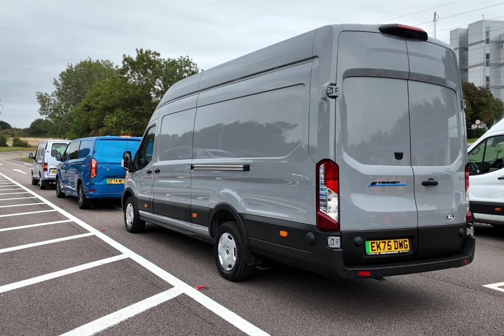 Szary samochód dostawczy typu van zaparkowany na drodze obok innych pojazdów w podobnej linii, w tle drzewa oraz fragment budynku przemysłowego.