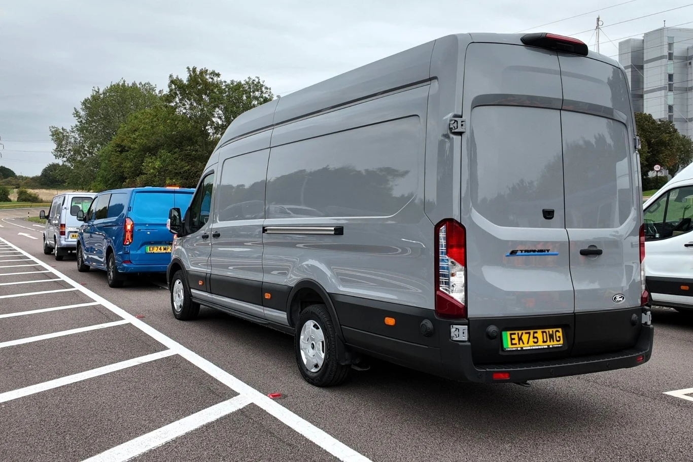 Szary samochód dostawczy typu van zaparkowany na drodze obok innych pojazdów w podobnej linii, w tle drzewa oraz fragment budynku przemysłowego.