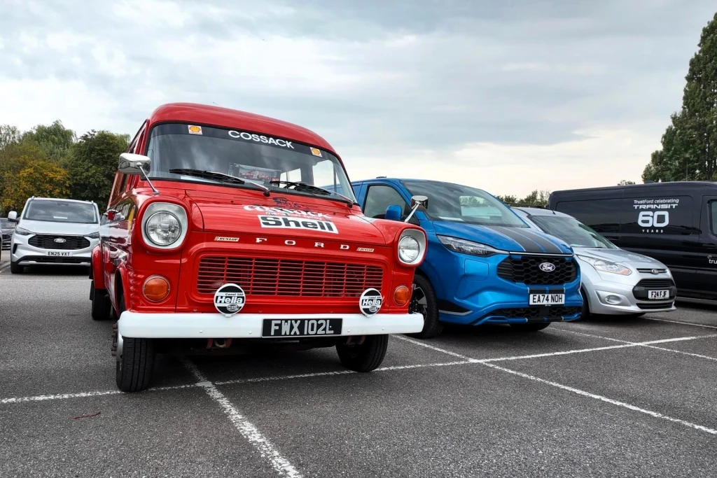 Czerwony klasyczny van Ford z logo Shell na masce, zaparkowany obok nowoczesnych samochodów osobowych, na asfaltowym parkingu przy pochmurnym niebie.