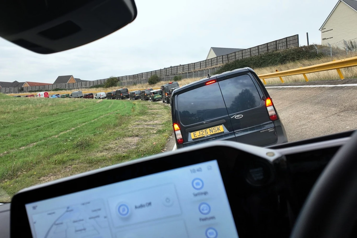 Kolumna samochodów poruszająca się wzdłuż drogi na lekko pagórkowatym terenie, widok z wnętrza pojazdu na dashboard i tylne światła czarnego vana z brytyjską tablicą rejestracyjną, w tle widać ogrodzenie i domy mieszkalne.