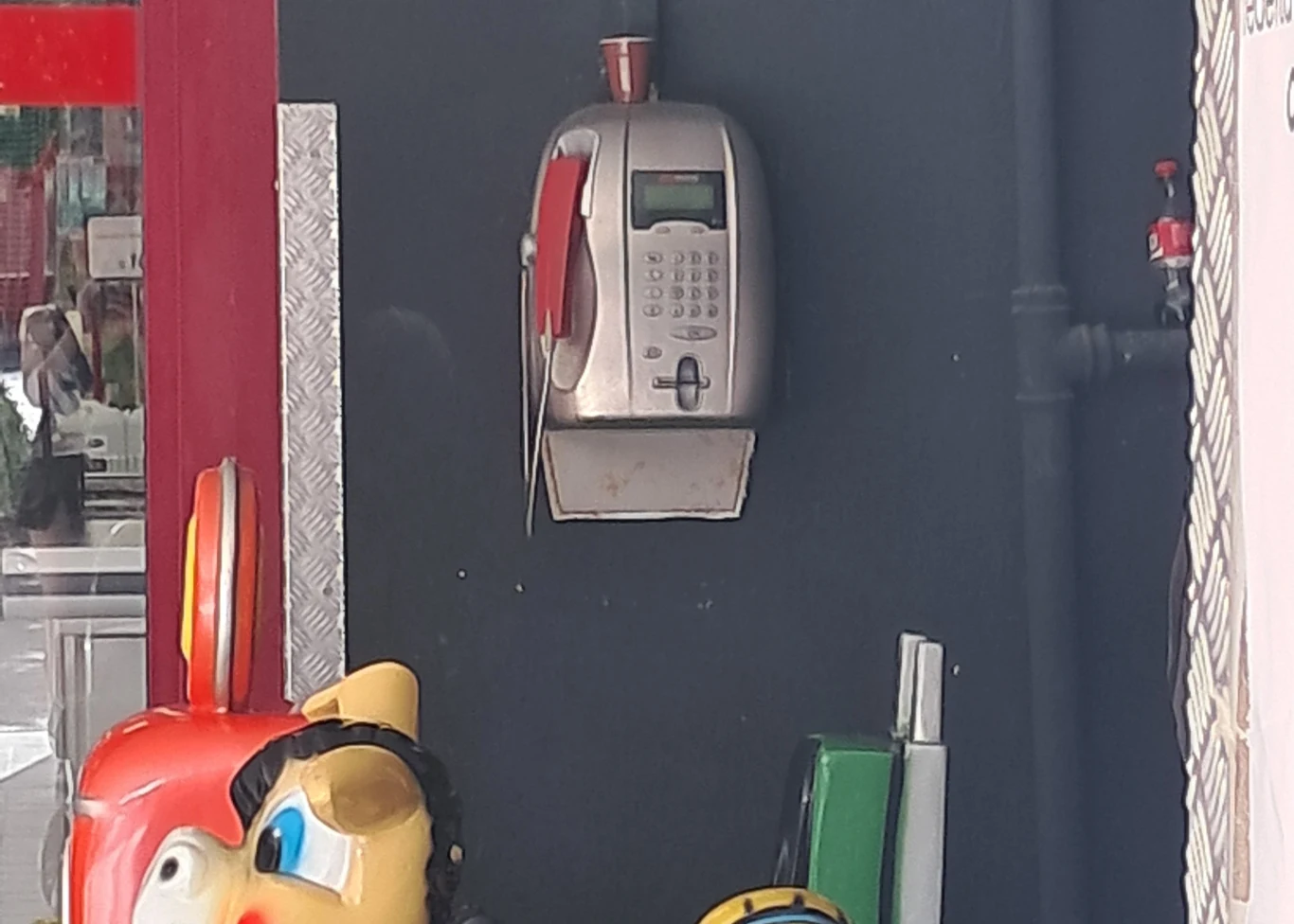 Telefon publiczny zamontowany na ścianie wraz z kolorowym automatem do zabawy dla dzieci częściowo widocznym na pierwszym planie.