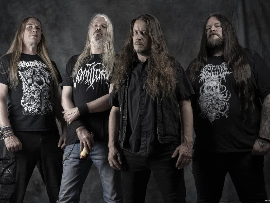 Asphyx Czterech mężczyzn z długimi włosami stoi obok siebie na ciemnym tle, ubrani w czarne koszulki z motywami death metalowymi, jeden z nich trzyma butelkę alkoholu.