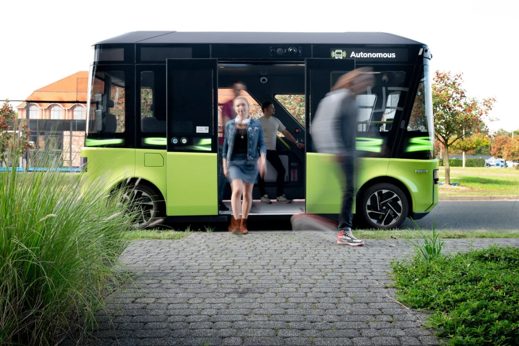 Ludzie wychodzący z autonomicznego pojazdu komunikacji miejskiej o nowoczesnym wyglądzie, zaparkowanego na ulicy w spokojnej okolicy, w tle widoczne rośliny i budynki.