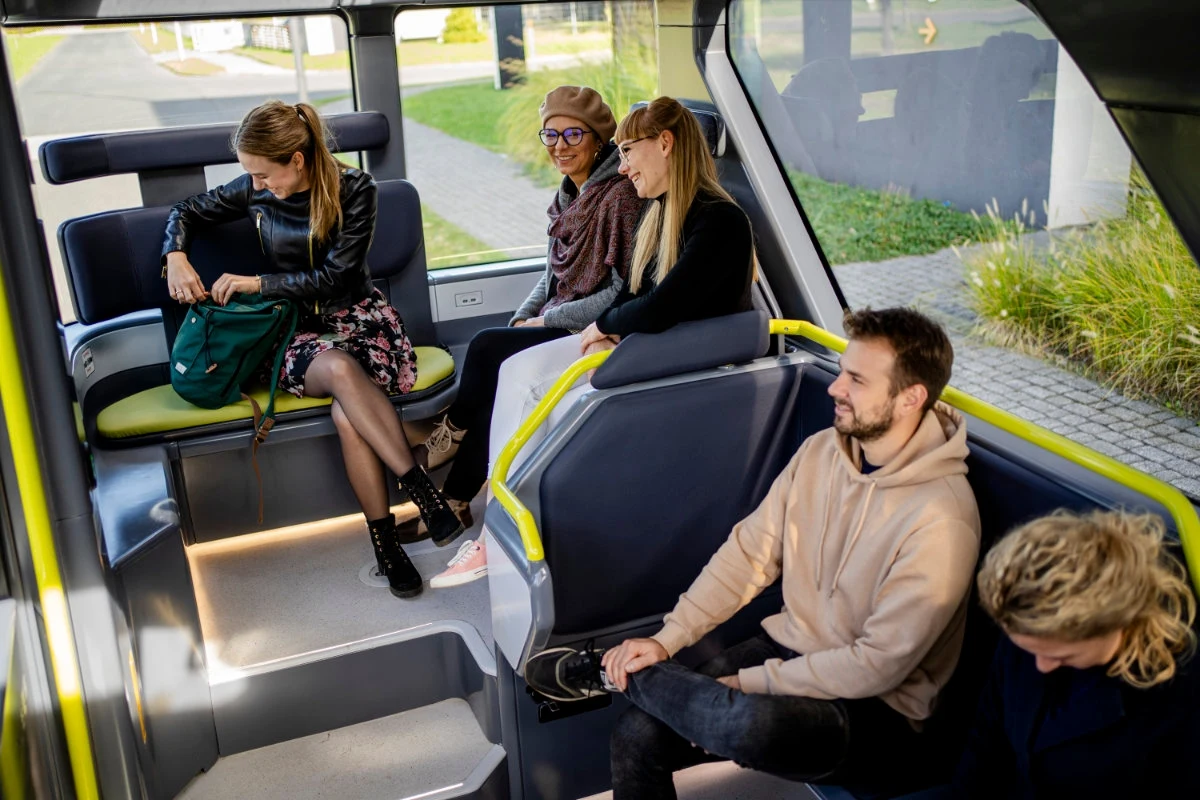 Grupa młodych osób siedzi w nowoczesnym, przestronnym wnętrzu pojazdu komunikacji miejskiej, zwracając się do siebie, rozmawiając i relaksując w jasnym, przyjemnym otoczeniu.