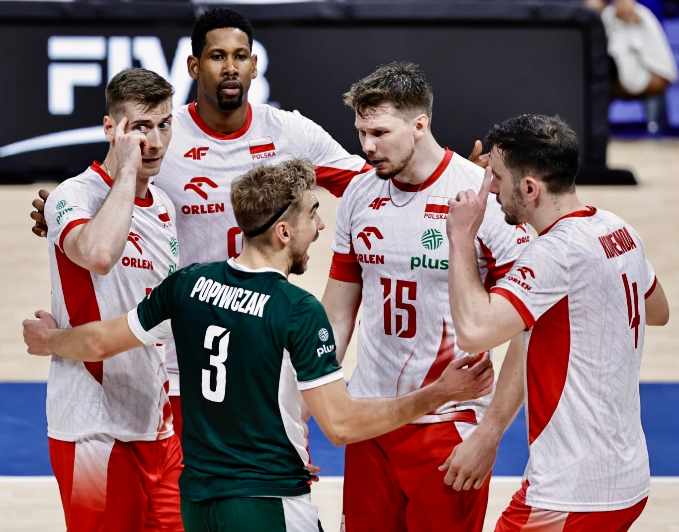 Grupa polskich siatkarzy podczas żywej wymiany zdań na boisku, w centrum znajduje się zawodnik w zielonej koszulce z numerem 3 Popiwczak, otoczony przez czterech kolegów w biało-czerwonych strojach, na tle hali sportowej.