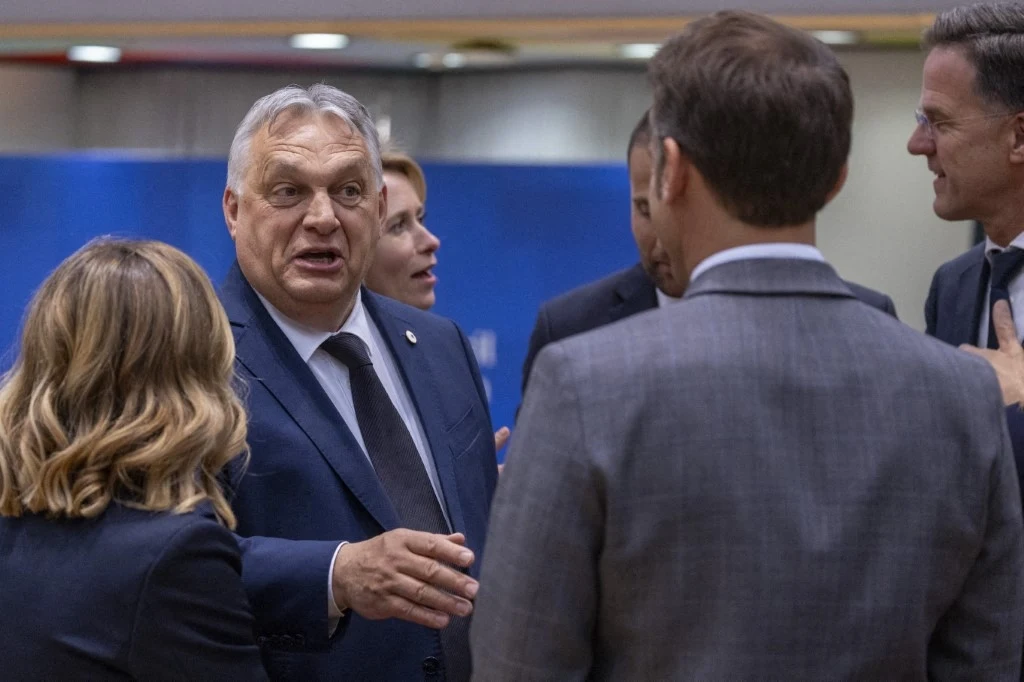 Viktor Orban rozmawiający z Emmanuelem Macronem podczas szczytu UE Kilka osób w garniturach prowadzi rozmowę w formalnej atmosferze, jedna z osób gestykuluje ręką, wyrażając emocje podczas dyskusji.