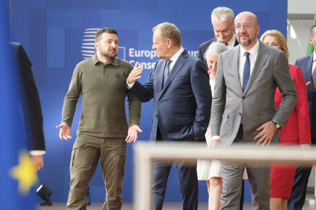 Wołodymyr Zełenski, Donald Tusk i Charles Michel przed szczytem UE w Brukseli Grupa polityków w garniturach i jeden mężczyzna w wojskowej koszulce rozmawiają na tle ściany z logo instytucji europejskiej, atmosfera profesjonalna, dyskusja w trakcie oficjalnego wydarzenia.