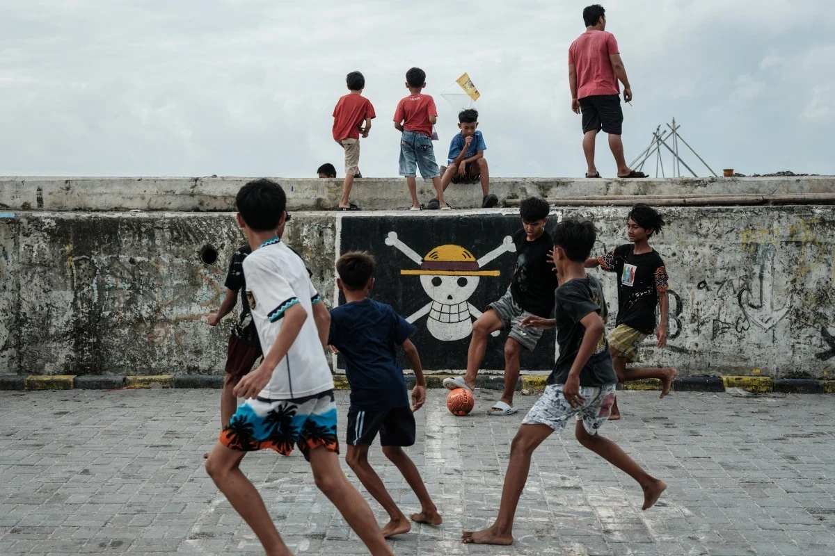 Flaga z One Piece wywieszona w Indonezji Grupa chłopców grających w piłkę nożną na brukowanym placu, w tle mur ozdobiony kolorowym graffiti z motywem pirata i czaszki.