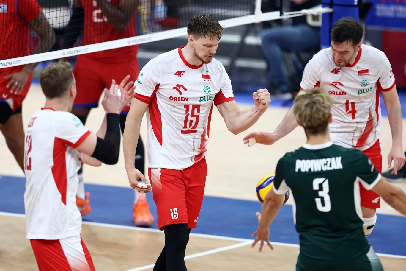 Jakub Kochanowski (nr 15) Siatkarze drużyny Polski w trakcie meczu na hali sportowej świętują zdobycie punktu, w tle widoczna siatka oraz zawodnicy przeciwnej drużyny.