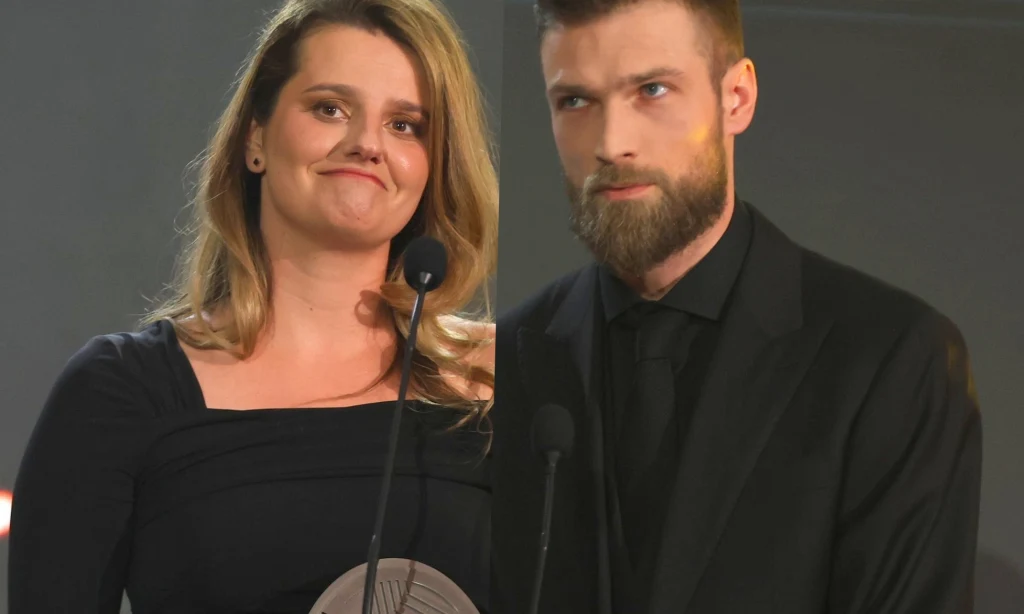 Zofia Zborowska i Andrzej Wrona Dwie osoby stoją przy mikrofonach, elegancko ubrane, kobieta z delikatnym uśmiechem i mężczyzna o poważnym wyrazie twarzy, jednolite tło w odcieniach szarości.