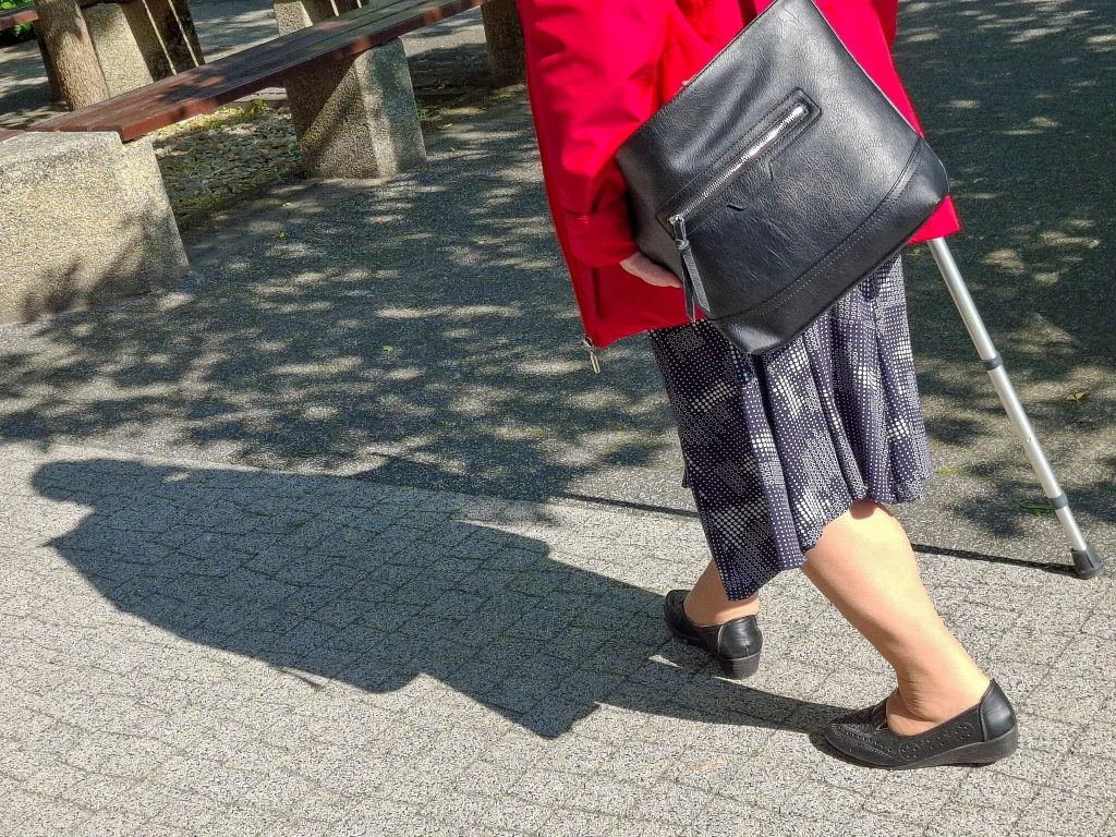 Kobieta w czerwonym płaszczu idąca z laską po chodniku, ubrana w ciemną spódnicę i czarne buty, z czarną torebką na ramieniu, rzuca cień na chodnik w słoneczny dzień.