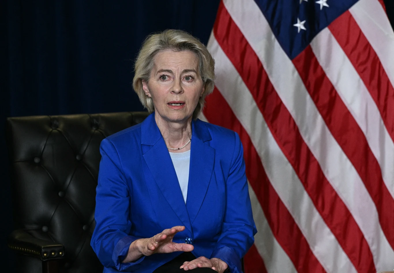 Komisja Europejska zapowiada przedstawienie propozycji ceł na rosyjską ropę Kobieta w niebieskiej marynarce siedzi na czarnym skórzanym fotelu na tle dużej flagi Stanów Zjednoczonych, wykonując gest ręką.