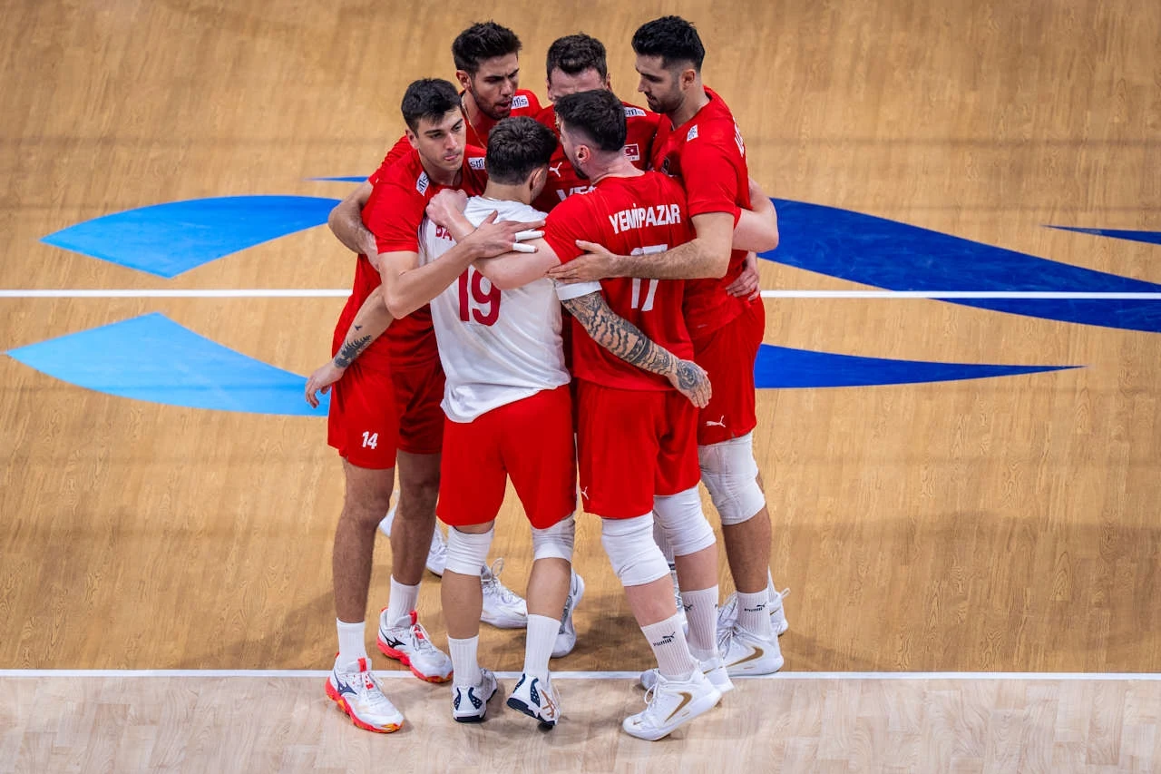 Grupa siatkarzy w czerwonych strojach świętująca udaną akcję, obejmując się na środku boiska o drewnianej nawierzchni.